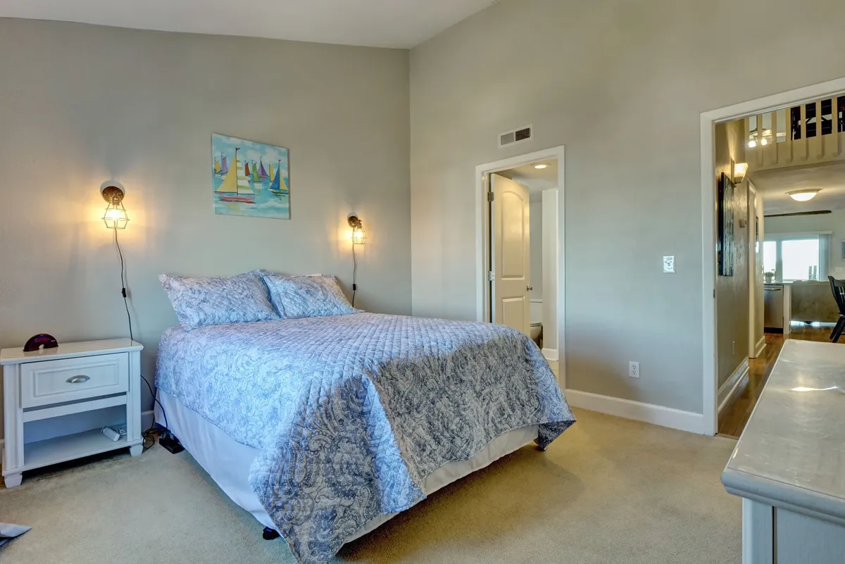 A bright, inviting bedroom scene with a bed covered in a blue quilt, complemented by wall art and a nightstand with a glowing lamp. Soft lighting and carefully chosen decor create a peaceful and intimate setting.