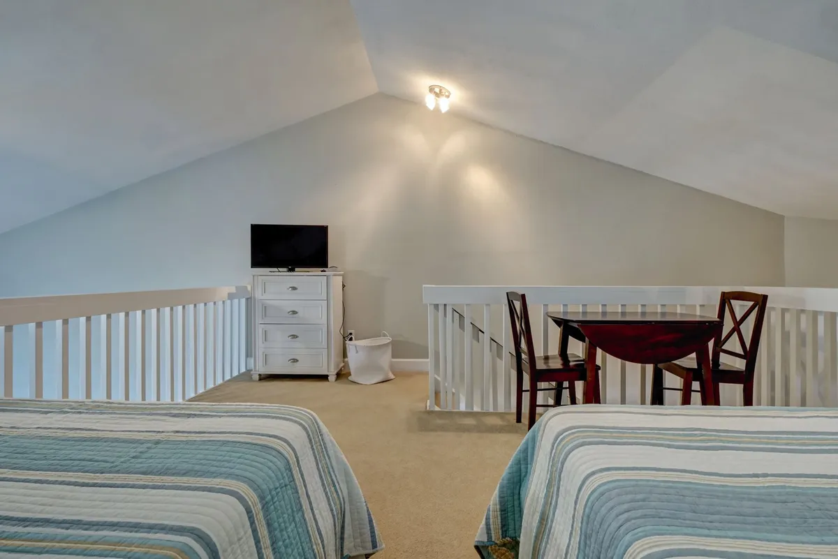 A bright, inviting loft bedroom featuring two full-sized beds with coordinated striped bedding. A small dining table with chairs sits nearby, and a television rests atop a dresser at the foot of one bed. Sunlight streams through a window, creating an airy, cheerful atmosphere ideal for family relaxation or playful sleepovers.