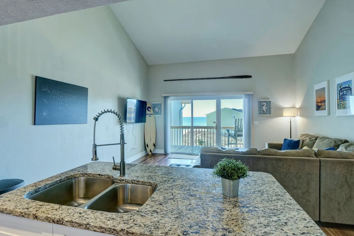 A bright, open-plan living area featuring a spacious kitchen with a granite island, white cabinetry, and stainless steel appliances. Large windows and a comfortable seating area offer views of the ocean, while a surfboard decoration adds a beachy touch and a TV hangs on the wall. The room is flooded with natural light, creating a welcoming and serene atmosphere.