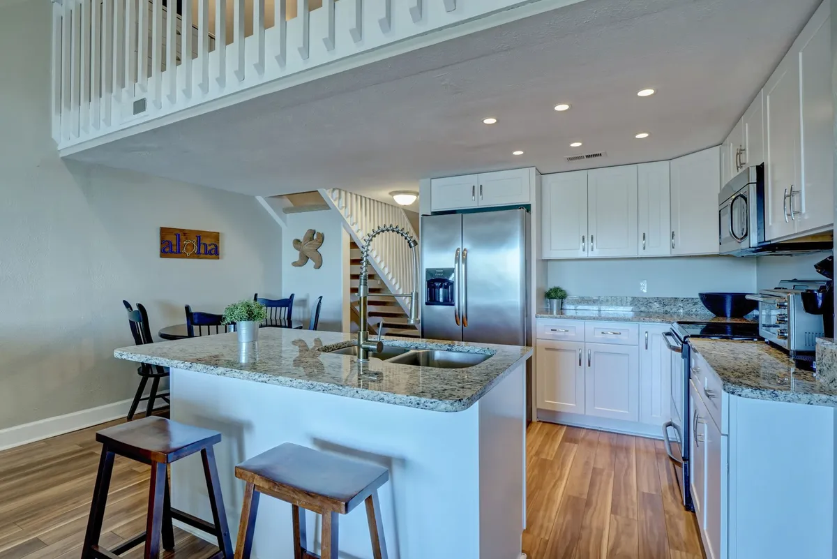 A bright, updated kitchen with granite countertops, stainless steel appliances, white cabinetry, and a staircase visible in the background. Decorative wall art adds personality, and the space feels airy and cheerful—ideal for preparing meals after a day spent exploring outside.