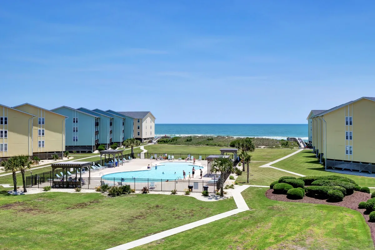 A bright, inviting swimming pool with clear blue water is surrounded by sun loungers and palm trees. In the background, the ocean's horizon is visible, along with the condominium buildings framing the pool area. The scene is active yet relaxed, suggesting a fun and family-friendly atmosphere.