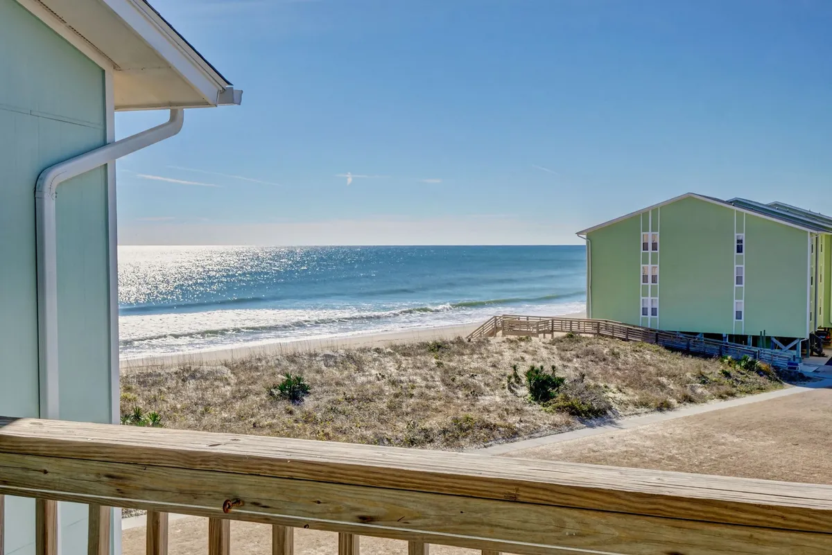 The hero image showcases an expansive ocean view with a pristine sandy beach stretching out in front, complemented by a distinctive green building—presumably the condo complex itself. Gentle waves lap the shore, inviting guests to stroll, sunbathe, or simply relax. The tranquil mood is underscored by sunrise or sunset lighting, suggesting peaceful mornings and memorable evenings by the sea.