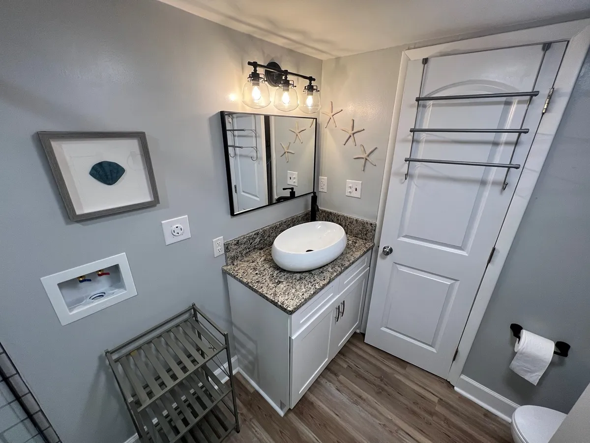 A modern en-suite bathroom featuring a granite countertop with an oval sink, a large mirror with ample lighting, starfish decor, and a storage rack. The calming decor and functionality suggest a space designed for self-care and relaxation.