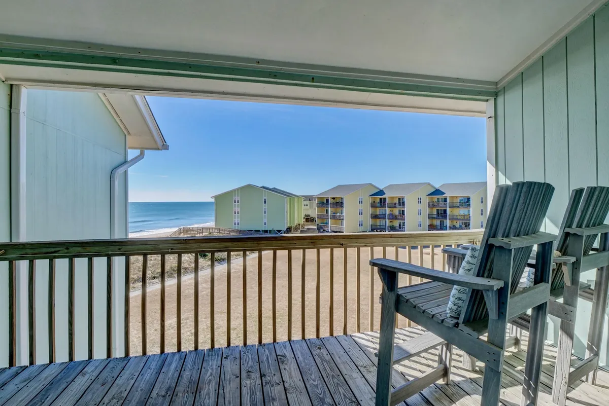 A serene wooden deck with comfortable chairs facing the ocean, set for relaxing and enjoying the view. The beach and water are visible beyond the deck, with colorful buildings adding charm to the coastal setting.