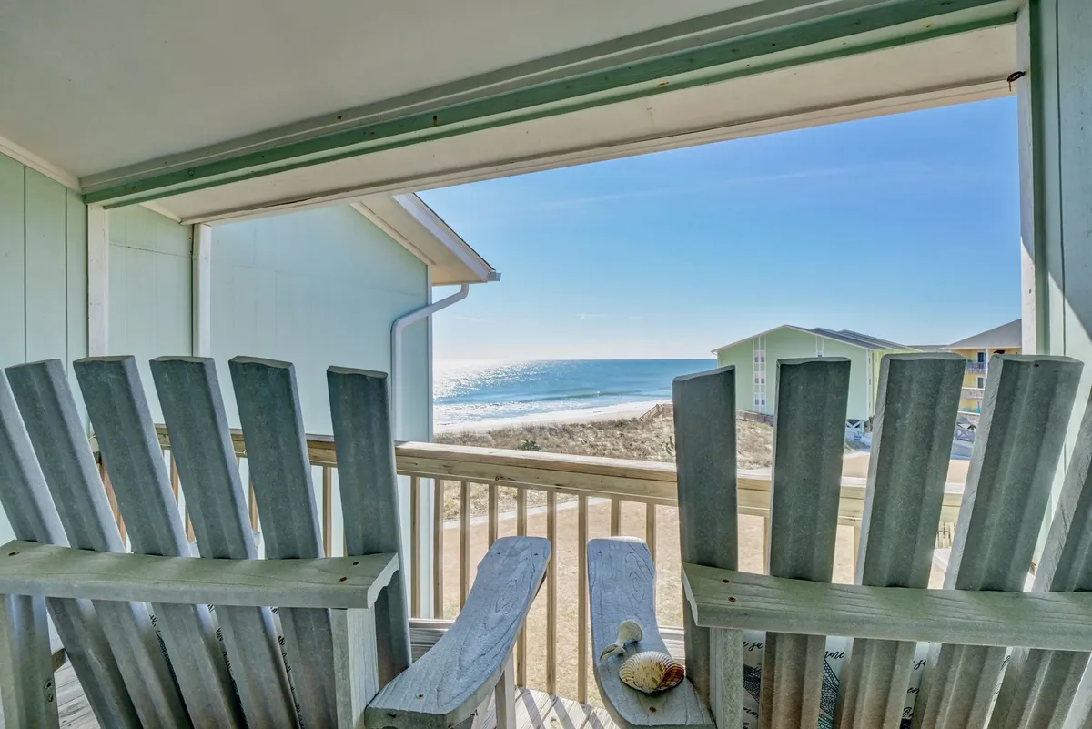 A pair of Adirondack chairs on a private deck overlook the ocean, with a shell placed on a small table between them. The scene is peaceful, evoking early mornings with coffee or tranquil evenings watching the waves.
