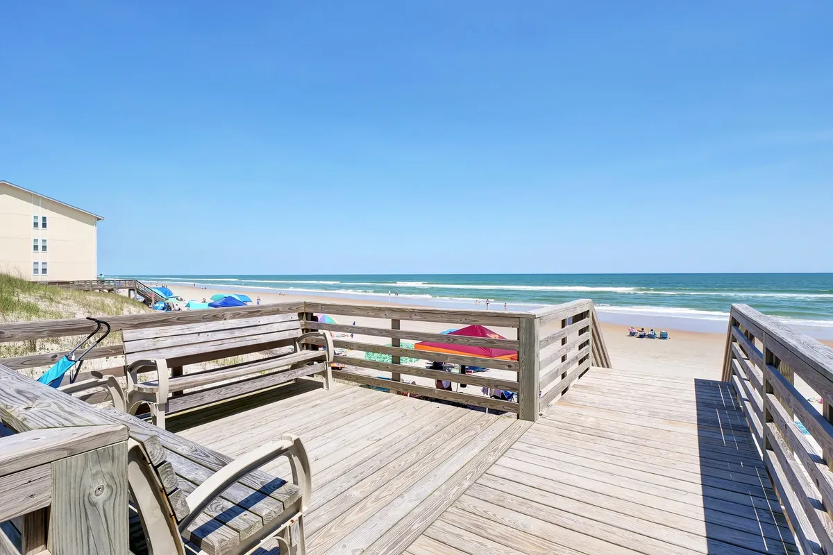 A spacious wooden deck with comfortable seating that opens onto a sandy beach and the wide ocean beyond. The scene is sunlit and inviting, suggesting opportunities for gatherings, picnics, and easy access to waterfront activities.