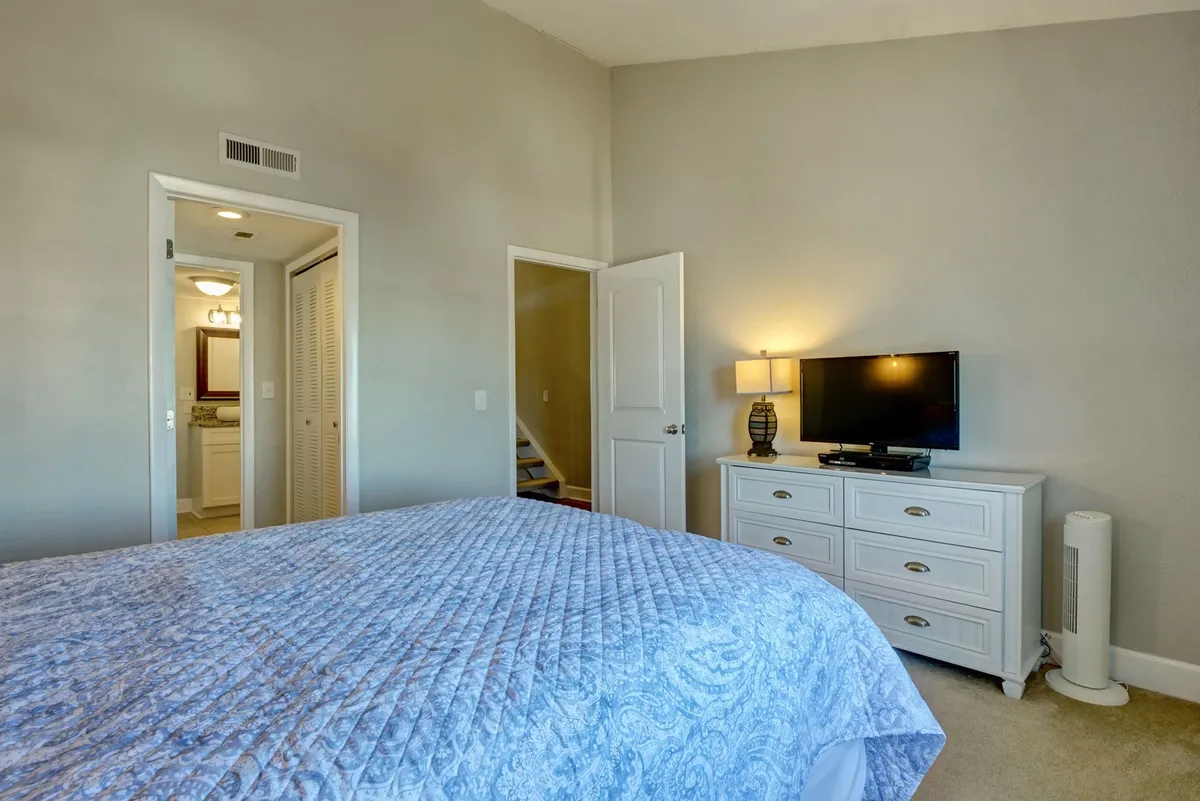 This image showcases a master bedroom featuring a plush bed with a blue quilt, a white dresser, a lamp on the bedside table, and a television. The arrangement exudes comfort, functionality, and a serene atmosphere ideal for relaxing after a busy day.
