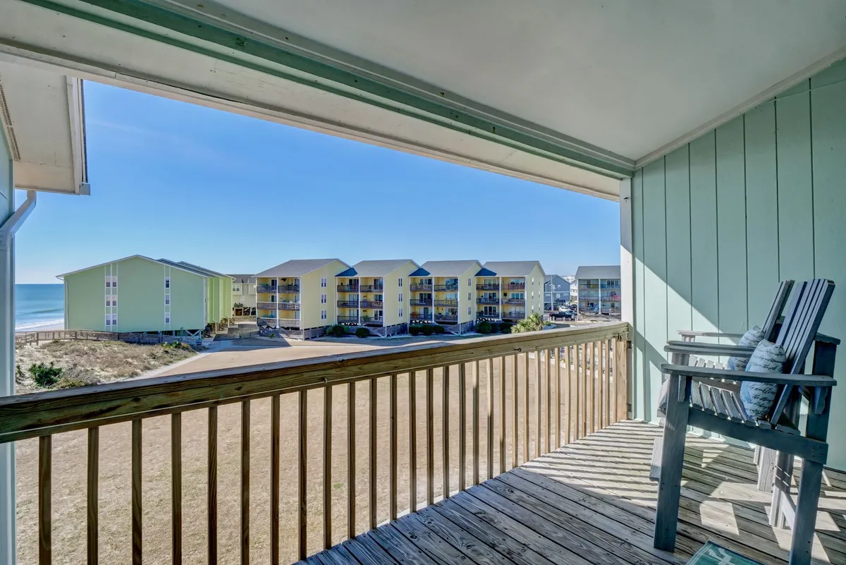 A tranquil balcony view overlooking the ocean, with a wooden railing in the foreground, green buildings below, and the surf gently meeting the sandy beach. The setting invites relaxation while staying connected to the vibrant coastal environment.