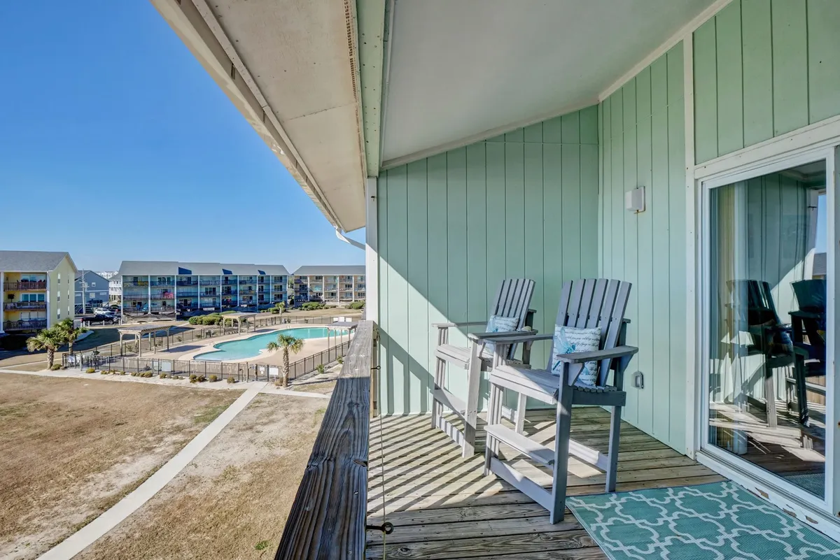 A tranquil view from a condo balcony featuring two wooden chairs overlooking the pool area below. The green building facade and lush landscaping set a peaceful atmosphere, inviting guests to imagine relaxing after a swim or socializing outdoors.