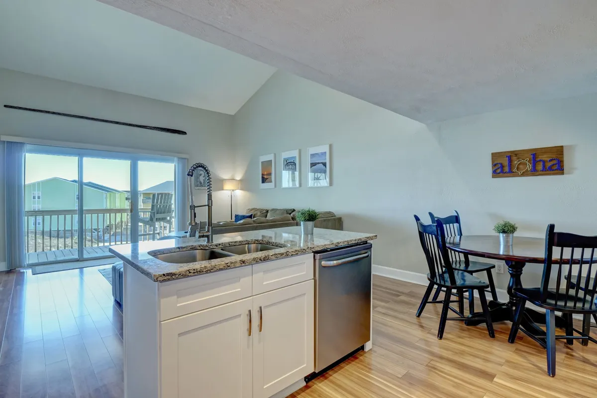 A view from the kitchen island, featuring a sink, adjacent dining table, and sliding glass door leading directly to the covered deck outside. Decor details like a cheerful 'aloha' sign add personality. The scene is open, airy, and designed for communal living.