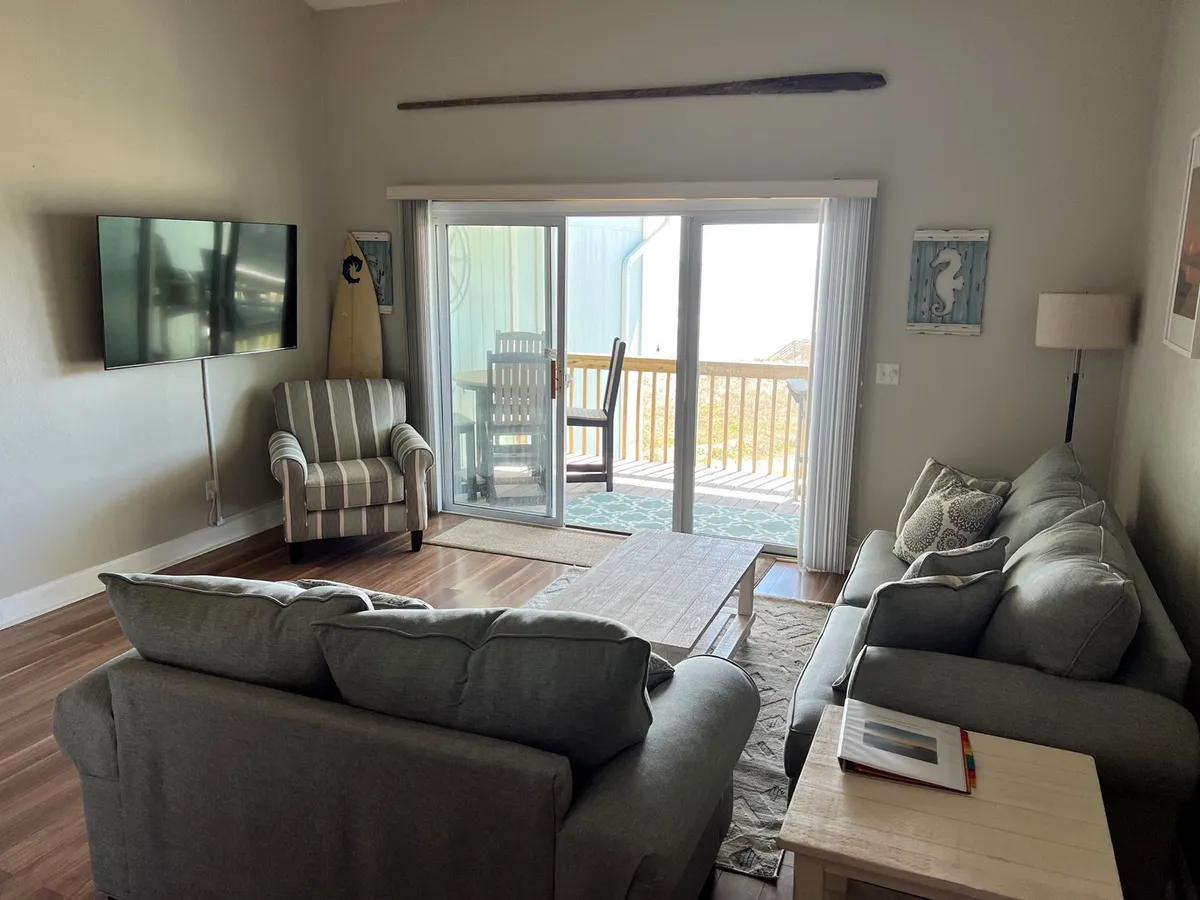 A cozy living room setup with a plush sofa facing a television. Sliding glass doors provide access to the deck and allow sunlight to pour in, offering a glimpse of the outdoor space and the ocean beyond.
