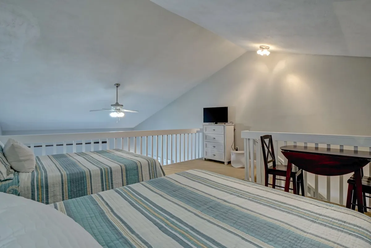 A spacious loft area with two beds dressed in striped bedding, a small dining table with chairs, and a dresser with a television. The room feels cozy yet open, illuminated by natural light that enhances the welcoming vibe.