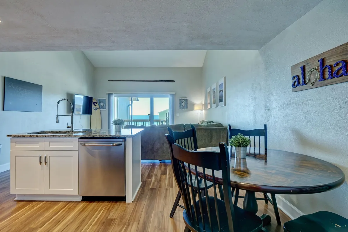 A kitchen and dining space featuring a kitchen island, dining table, a large window letting in abundant natural light, and a decorative 'aloha' sign. The window gives a sense of connection to the outdoors, and the layout suggests a welcoming area for returning from island adventures, preparing meals, and gathering.