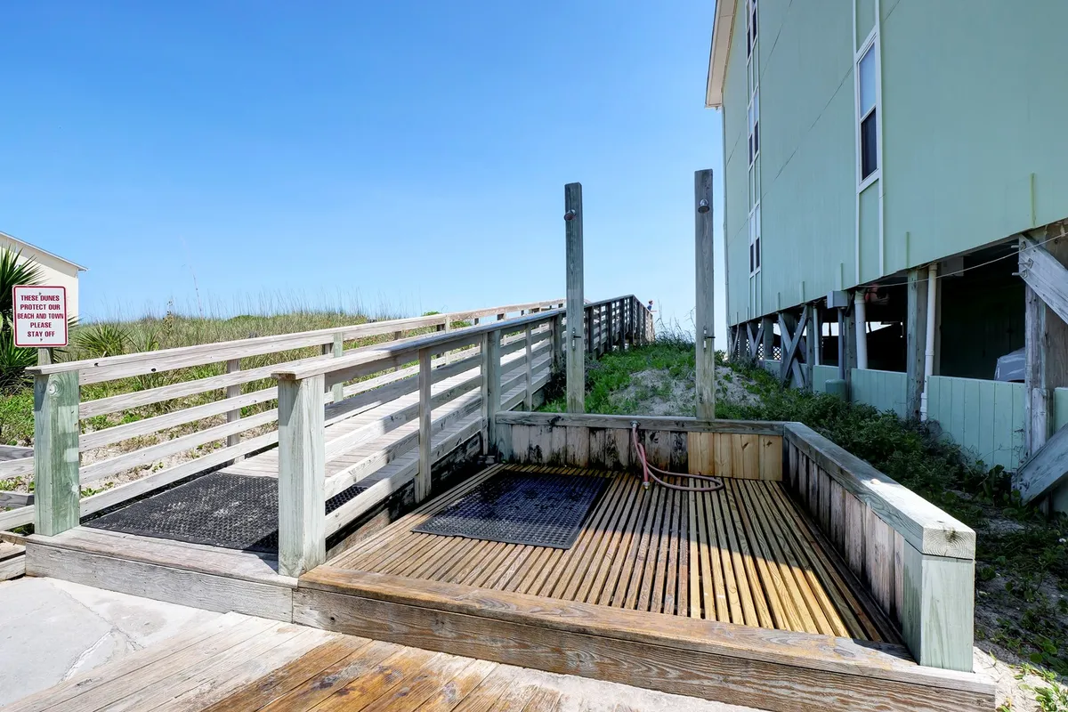 A wooden deck leading to a dedicated outdoor shower and a pathway that continues directly to the sandy beach. The adjacent green building is visible, suggesting proximity to the condo units. The setting is sunlit and laid-back, emphasizing convenience and a seamless transition from beach to home.