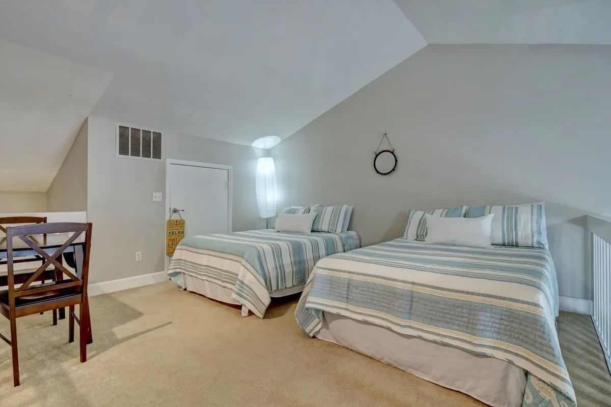 An inviting interior space with two beds featuring striped bedding, a small dining table with chairs, and a decorative wall mirror. The arrangement encourages relaxation, conversation, and shared moments, with high ceilings contributing to an airy atmosphere.